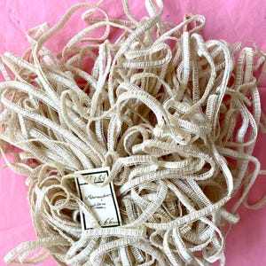 May include: A mass of off-white, textured ribbon, possibly vintage, on a pink background. A small tag with handwritten text is attached. The ribbon is likely for sewing or craft projects. The ribbon is in a tangled pile.