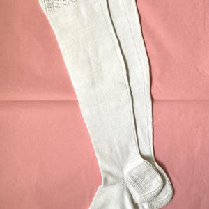 Puede incluir: Un par de medias blancas hasta la rodilla con un delicado puño de punto de encaje en la parte superior. La tela tiene una textura finamente acanalada en toda su extensión, y una media muestra una pequeña letra S roja bordada cerca del borde.