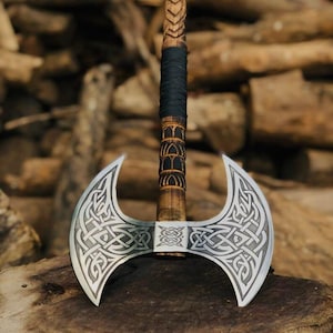 Puede incluir: Un hacha de doble hoja con un mango de madera y intrincados diseños de nudos celtas en las hojas plateadas. El mango está envuelto en cuero negro en la parte superior.