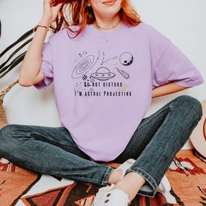 Op de afbeelding: Een lichtpaars T-shirt met een zwarte afbeelding van een melkwegstelsel, planeten en een UFO. De tekst op het shirt luidt "DO NOT DISTURB I'M ASTRAL PROJECTING". Het shirt wordt gedragen met een blauwe jeans en witte sneakers.