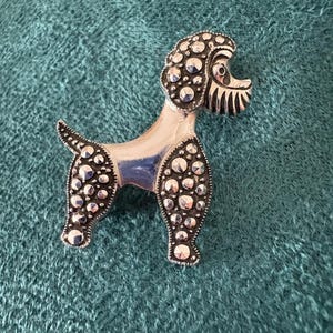 Puede incluir: Broche de caniche plateado con superficie texturizada. El cuerpo y la cabeza del caniche están adornados con pequeños acentos redondos plateados. El broche está sobre un fondo de tela color verde azulado.