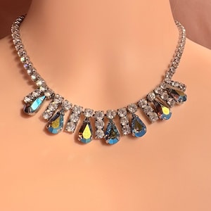 May include: A silver-toned necklace with clear, round rhinestones and iridescent teardrop-shaped pendants. The necklace is displayed on a mannequin, showcasing its design and sparkle. The teardrop pendants have a blue and gold color.