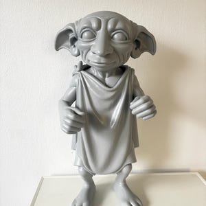 Könnte beinhalten: Graue Figur einer fiktiven Figur mit großen Ohren, einer markanten Nase und großen Augen. Die Figur trägt ein drapiertes Gewand und hat einen detaillierten Gesichtsausdruck. Die Figur steht auf einer weißen Oberfläche.