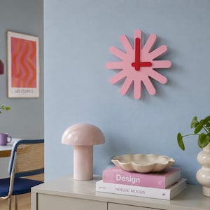Könnte beinhalten: Eine rosa Wanduhr im Sonnenstrahl-Stil mit roten Zeigern hängt an einer hellblauen Wand. Eine rosa pilzförmige Lampe, eine weiße, wellenförmige Schale und Bücher stehen auf einem grauen Schrank. Eine Vase mit Grünzeug steht ebenfalls auf dem Schrank.