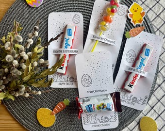 Puede incluir: Un arreglo temático de Pascua con barras de chocolate Kinder, caramelos y tarjetas decorativas. Las tarjetas presentan ilustraciones de huevos y conejos de Pascua, con textos como "Felices Pascuas". Una ramita de sauce y decoraciones de Pascua completan la escena.