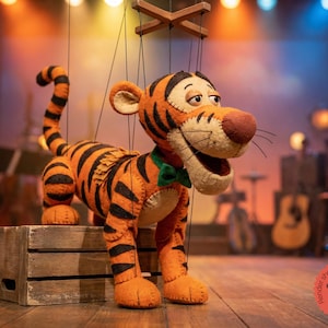 Op de afbeelding: Een handgemaakte tijgerpop met oranje en zwarte strepen, een groene vlinderdas en een glimlachende uitdrukking. De pop hangt aan touwtjes en staat op een houten kist. De pop is ongeveer 61 cm hoog.