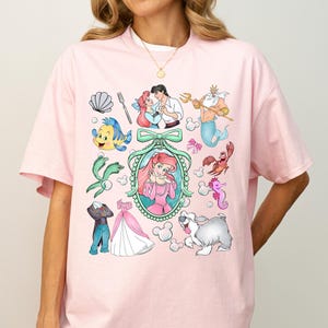 Pu&ograve; includere: Una maglietta rosa chiaro con una grafica colorata di personaggi dei cartoni animati. Il design include una sirena, un principe e altre creature marine. La maglietta ha maniche corte e scollo tondo.