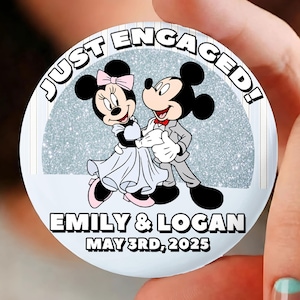 May include: A round button with the text "JUST ENGAGED!" featuring a cartoon couple, Minnie and Mickey Mouse. The button includes the names "EMILY & LOGAN" and the date "MAY 3RD, 2025" in white text on a light blue background.