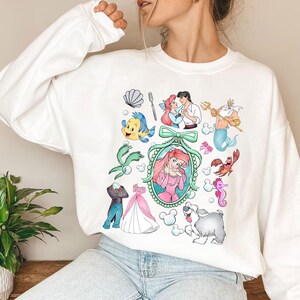 Pu&ograve; includere: Felpa bianca con illustrazioni colorate di personaggi ed elementi di un popolare film d'animazione. Il design include Ariel, Flounder, Sebastian, il Principe Eric, Re Tritone e altre immagini iconiche. Un capo d'abbigliamento comodo e casual.