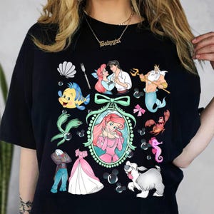 Pu&ograve; includere: T-shirt nero con un design a cartoni animati colorati con personaggi di un popolare film d'animazione. Il design include una sirena, un pesce e un granchio, insieme ad altre immagini correlate. La maglietta ha uno scollo tondo.