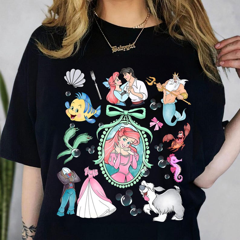 Pu&ograve; includere: T-shirt nero con un design a cartoni animati colorati con personaggi di un popolare film d'animazione. Il design include una sirena, un pesce e un granchio, insieme ad altre immagini correlate. La maglietta ha uno scollo tondo.