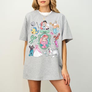 Pu&ograve; includere: Una maglietta grigia con una grafica colorata di personaggi ed elementi di un popolare film d'animazione. Il design include un ritratto centrale, un vestito e varie creature marine. La maglietta &egrave; indossata da una persona.