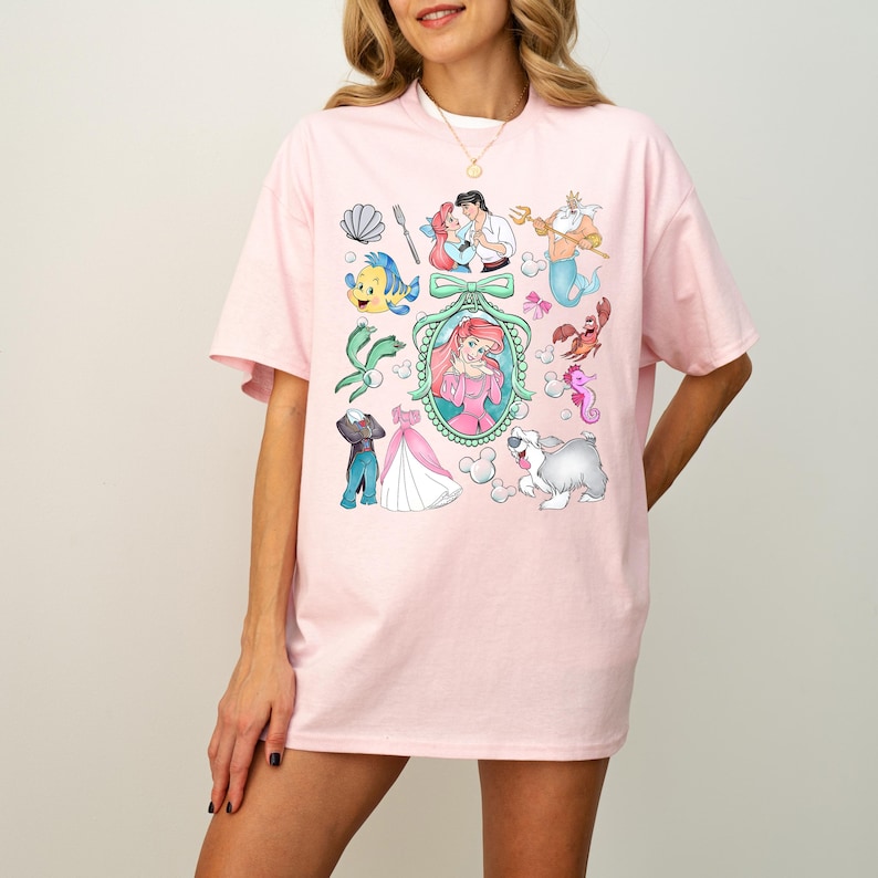 Pu&ograve; includere: Una maglietta rosa chiaro con una grafica colorata di personaggi dei cartoni animati. Il design include una sirena, un principe e altre creature marine. La maglietta ha maniche corte e scollo tondo.