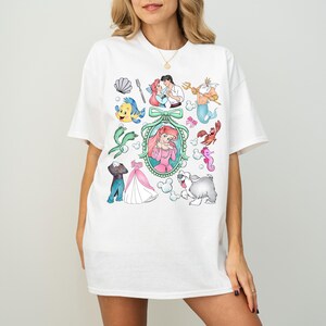 Pu&ograve; includere: T-shirt bianco con illustrazioni di personaggi dei cartoni animati di un popolare film d'animazione. Il design include una sirena, un granchio, un pesce e altri personaggi. La maglietta ha una vestibilit&agrave; casual e oversize.