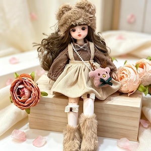May include: A doll with long brown hair wearing a fuzzy brown bear-ear hat, a brown cable-knit sweater with a small bow tie, and a beige corduroy overall skirt. The doll carries a small pink plush deer bag and wears white socks with bows and fuzzy boots.