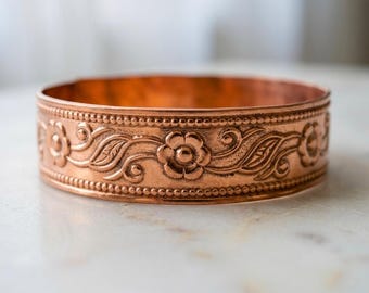 Pode incluir: Um bracelete de metal na cor cobre com um padrão em relevo contínuo de flores de cinco pétalas e motivos de vinhas, contornado por duas fileiras finas de detalhes de contas em relevo ao longo das bordas superior e inferior da peça.