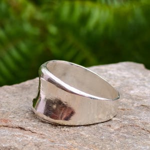 Peut inclure: Une large bague en argent poli avec une surface lisse et incurvée et un profil légèrement concave. L'anneau présente une finition métallique réfléchissante qui capte la lumière, montrant une construction solide et continue sans gravures.