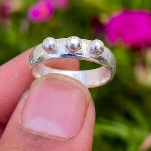 Puede incluir: Un anillo de plata pulida con una banda lisa y redondeada, adornado con tres pequeñas cuentas esféricas espaciadas uniformemente a lo largo del borde superior. El metal tiene un acabado metálico reflectante que capta la luz.