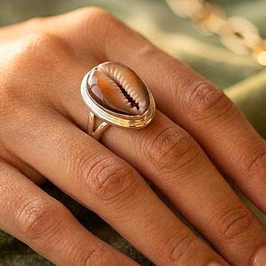 May include: A gold ring with a large, oval-shaped shell as its centrepiece. The shell displays brown and cream hues and intricate detailing. The ring is worn on a hand with manicured nails, with a gold chain visible in the background.