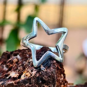 Puede incluir: Anillo plateado con forma de estrella de cinco puntas. El anillo se encuentra sobre una superficie texturizada y marrón, con un fondo borroso de hojas verdes.