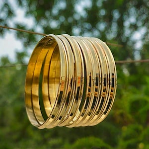 Può includere: Un set di molteplici bracciali rigidi sottili in metallo color oro lucido impilati insieme. Ogni bracciale presenta una superficie liscia e riflettente con un profilo arrotondato, creando un aspetto uniforme nell'intera collezione di bracciali circolari.