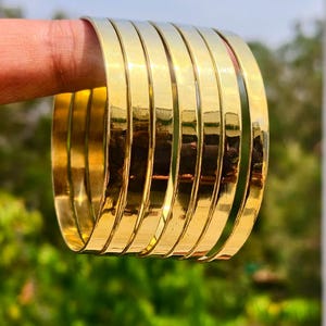 Puede incluir: Un conjunto de siete brazaletes de metal en tono dorado pulido con una superficie lisa y reflectante. Las pulseras circulares están apiladas, presentando un diseño simple y sin adornos con un ancho constante y un acabado amarillo metálico brillante.