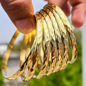 Peut inclure: Un ensemble de sept bracelets joncs en métal doré présentant une surface martelée et texturée. Chaque bracelet circulaire possède une finition polie et réfléchissante qui capte la lumière, créant un motif répété de petites empreintes concaves.