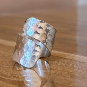 Op de afbeelding: Een zilveren ring met een gehamerde textuur. De ring heeft een wikkelontwerp, met twee overlappende banden. Het metaal heeft een glanzend, reflecterend oppervlak. De ring wordt gepresenteerd op een houten oppervlak.
