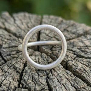 Op de afbeelding: Een zilveren ring met een cirkelvormig ontwerp. De ring heeft een dikke, matte band die een complete cirkel vormt. De ring is geplaatst tegen een getextureerd, verweerd houten oppervlak, met een vage groene achtergrond.