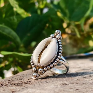 Op de afbeelding: Een zilveren ring met een grote, ovale, witte zeeschelp als middelpunt. De schelp is gezet in een gedetailleerde zilveren lijst met een kralenrand en decoratieve accenten. De ring wordt getoond op een houten oppervlak met een vage groene achtergrond.