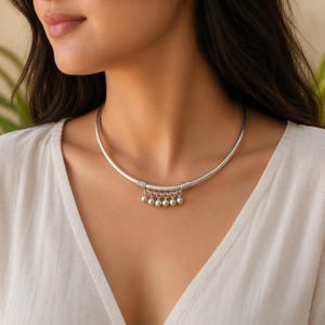 Könnte beinhalten: Eine silberfarbene Metall-Halskette in Choker-Form mit einem starren, glatten, kreisförmigen Band. Die Mitte der Kette ist mit fünf kleinen, hängenden, kugelförmigen Anhängern verziert, die an einzelnen Ringen befestigt und durch gewickelte Drahtdetails gesichert sind.