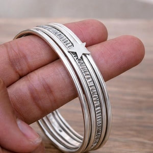 Peut inclure: Un ensemble de cinq bracelets joncs empilés de couleur argentée. Quatre bracelets ont une surface lisse et arrondie, tandis qu'un bracelet central présente un motif texturé à lignes verticales et une petite étoile plate à cinq branches.
