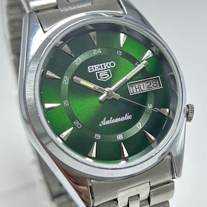 May include: A silver-tone stainless steel Seiko 5 automatic wristwatch with a sunburst green dial. The watch features silver hour markers, luminous hands, a day-date window at the three o'clock position, and a metal link bracelet strap.