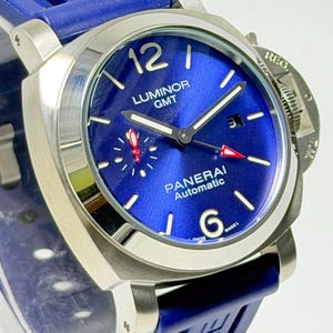 Könnte beinhalten: Eine Panerai Luminor GMT Automatikuhr mit einem sonnenschliff-blauen Zifferblatt, silberfarbenen Zeigern und arabischen Ziffern bei 12 und 6 Uhr. Die Uhr verfügt über ein Datumsfenster, ein kleines Sekunden-Hilfszifferblatt mit rotem Zeiger und ein blaues Kautschukarmband.