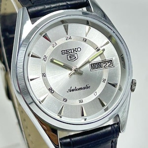 May include: A Seiko 5 automatic wristwatch with a silver sunray dial, silver-tone hour markers, and luminous hands. The watch features a day-date window at the three o'clock position and a black leather strap with a textured, crocodile-embossed finish.