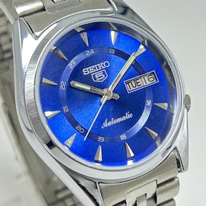 May include: A stainless steel Seiko 5 automatic wristwatch featuring a vibrant blue sunray dial with silver-tone hour markers, luminous hands, and a day-date window at the three o'clock position. The watch includes a matching metal link bracelet.
