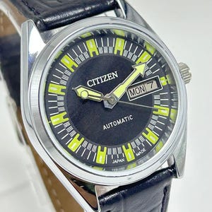 May include: A Citizen automatic wristwatch featuring a round silver-tone case and a black dial with bright yellow hour markers. The watch includes a day-date window at the three o'clock position and is attached to a black textured leather strap.