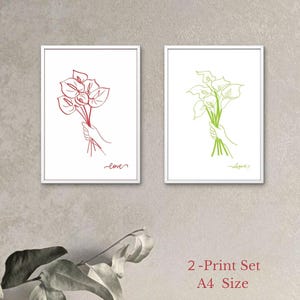 以下が含まれることがあります： 2つの額入りアートプリント。1つのプリントは、手に持った花の赤い線画で、下に「love」の文字があります。もう1つのプリントは、手に持った花の緑の線画で、下に「elegance」の文字があります。プリントはA4サイズです。