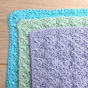 May include: Three crocheted washcloths in shades of blue, green, and lavender. Each cloth has a textured, floral-like pattern. The washcloths are stacked, with the blue one on the bottom, the green in the middle, and the lavender on top.