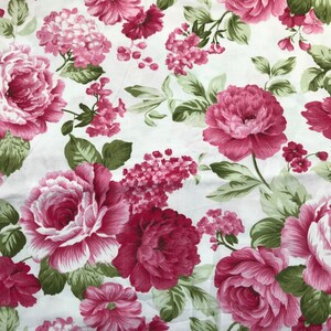 Könnte beinhalten: Weißer Stoff mit einem sich wiederholenden Blumenmuster. Große, offene Rosen in Rosa- und Burgundertönen sind mit kleineren Gruppen rosa Blüten und grünen Blättern durchsetzt. Ein klassisches, romantisches Design.