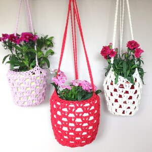 May include: Three hanging plant pots made of crocheted yarn in pink, red, and white. Each pot contains a green plant with pink flowers.