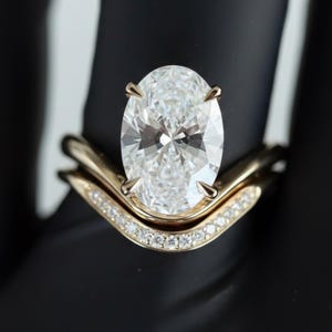 Könnte beinhalten: Ein goldener Verlobungsring-Set mit einem oval geschliffenen Diamanten und einem geschwungenen Band mit kleineren Akzentsteinen. Der Ring ist vor einem schwarzen Hintergrund platziert, um das Funkeln der Diamanten hervorzuheben. Der Markenname "Amella Jewels" ist sichtbar.