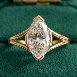 May include: A marquise-cut diamond ring with a gold band. The diamond is set in a gold bezel setting, and the band features a split shank design. The ring is displayed in a green velvet ring box.