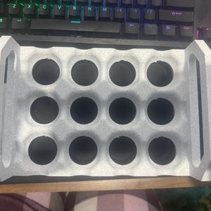 May include: A rectangular white speckled storage tray featuring twelve circular cutouts arranged in a three-by-four grid. The tray includes two integrated vertical handles on the short ends, providing a structured organiser for small items.