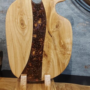Può includere: Tagliere in legno dal design unico. La tavola presenta legno di colore chiaro con un centro scuro riempito di resina. La resina è riempita di elementi decorativi. La tavola è sostenuta da un supporto in legno.