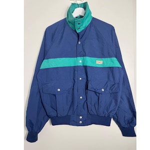 May include: A vintage blue jacket with a teal stripe across the chest and a teal collar. The jacket has two front pockets, a button-up closure, and ribbed cuffs. The jacket is hanging on a white hanger.