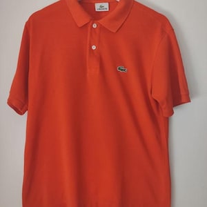 Könnte beinhalten: Leuchtend orangefarbenes Poloshirt mit kurzen Ärmeln, Kragen und einer Knopfleiste mit drei Knöpfen. Das Shirt hat ein kleines grünes Krokodil-Logo auf der Brust und ein weißes Etikett mit Text am Kragen. Es hängt an einem weißen Kleiderbügel.