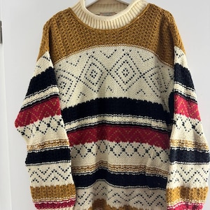 Peut inclure: Un pull surdimensionné avec un col roulé côtelé crème. Le pull présente un motif géométrique en crème, noir, rouge et or. Les manches et le corps ont des rayures horizontales. Le pull est suspendu à un cintre blanc.