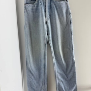 Könnte beinhalten: Hellblaue Jeans mit klassischem Fünf-Taschen-Design. Die Jeans hängen an einem schwarzen Kleiderbügel vor einer weißen Wand. Die Jeans haben einen Knopf- und Reißverschluss und eine gerade Beinpassform.