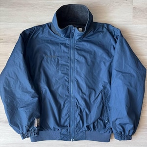 Op de afbeelding: Een marineblauwe jas met een hoge kraag en een ritssluiting over de hele lengte. De jas heeft een donkergrijze fleece voering op de kraag en manchetten. Een klein logo is zichtbaar op de linkerborst en een kleine patch op de linkermouw.